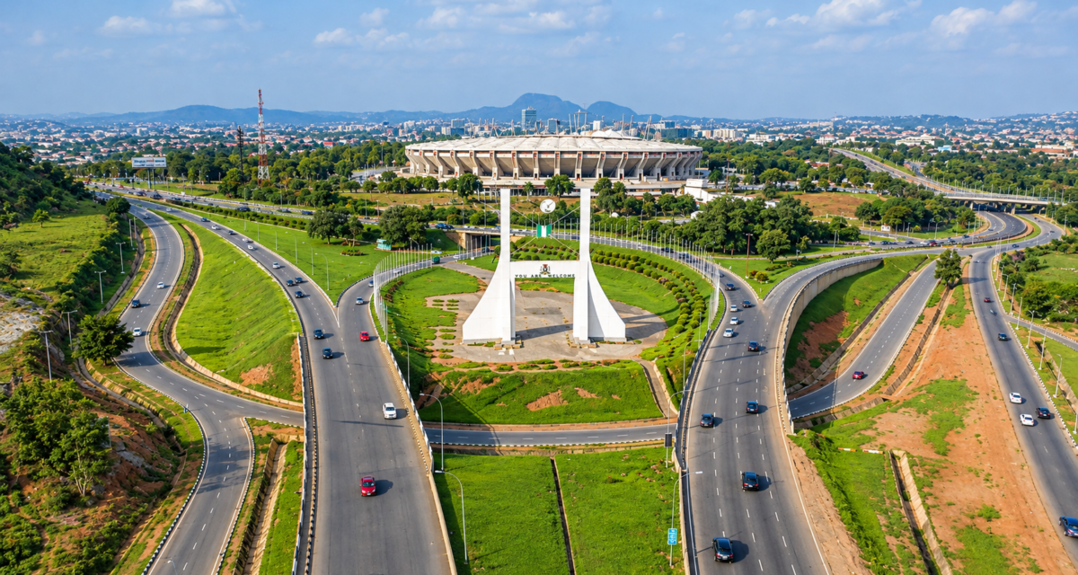 Aerial view of the Abuja City Gate monument surrounded by highway interchanges on Airport Road, with Moshood Abiola National Stadium visible in the background.