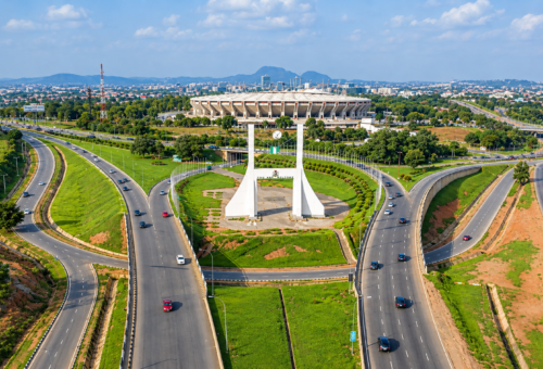Aerial view of the Abuja City Gate monument surrounded by highway interchanges on Airport Road, with Moshood Abiola National Stadium visible in the background.