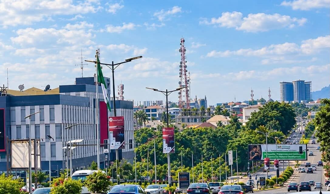 Ground-level view of a wide multi-lane road in Abuja with moderate car traffic, modern commercial buildings, streetlamps, and a clear blue sky.