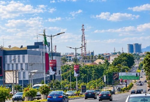 Ground-level view of a wide multi-lane road in Abuja with moderate car traffic, modern commercial buildings, streetlamps, and a clear blue sky.