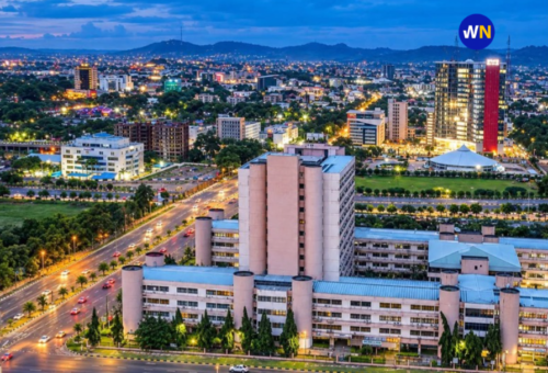 Elevated dusk cityscape of Abuja showing illuminated multi-lane expressways, modern government and commercial complexes, and sprawling glowing city lights extending toward distant hills.