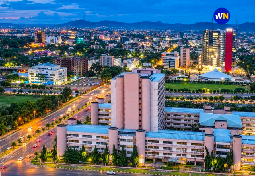 Elevated dusk cityscape of Abuja showing illuminated multi-lane expressways, modern government and commercial complexes, and sprawling glowing city lights extending toward distant hills.