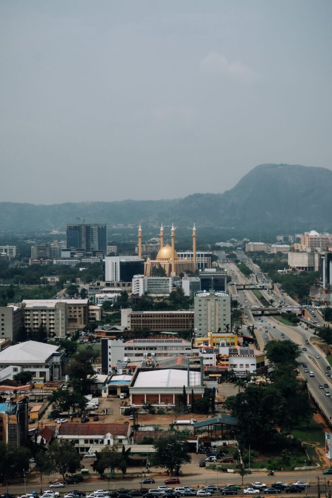 Elevated cityscape view of Abuja's Central Business District featuring the golden dome and minarets of the Nigerian National Mosque against rolling hills and wide roadways.